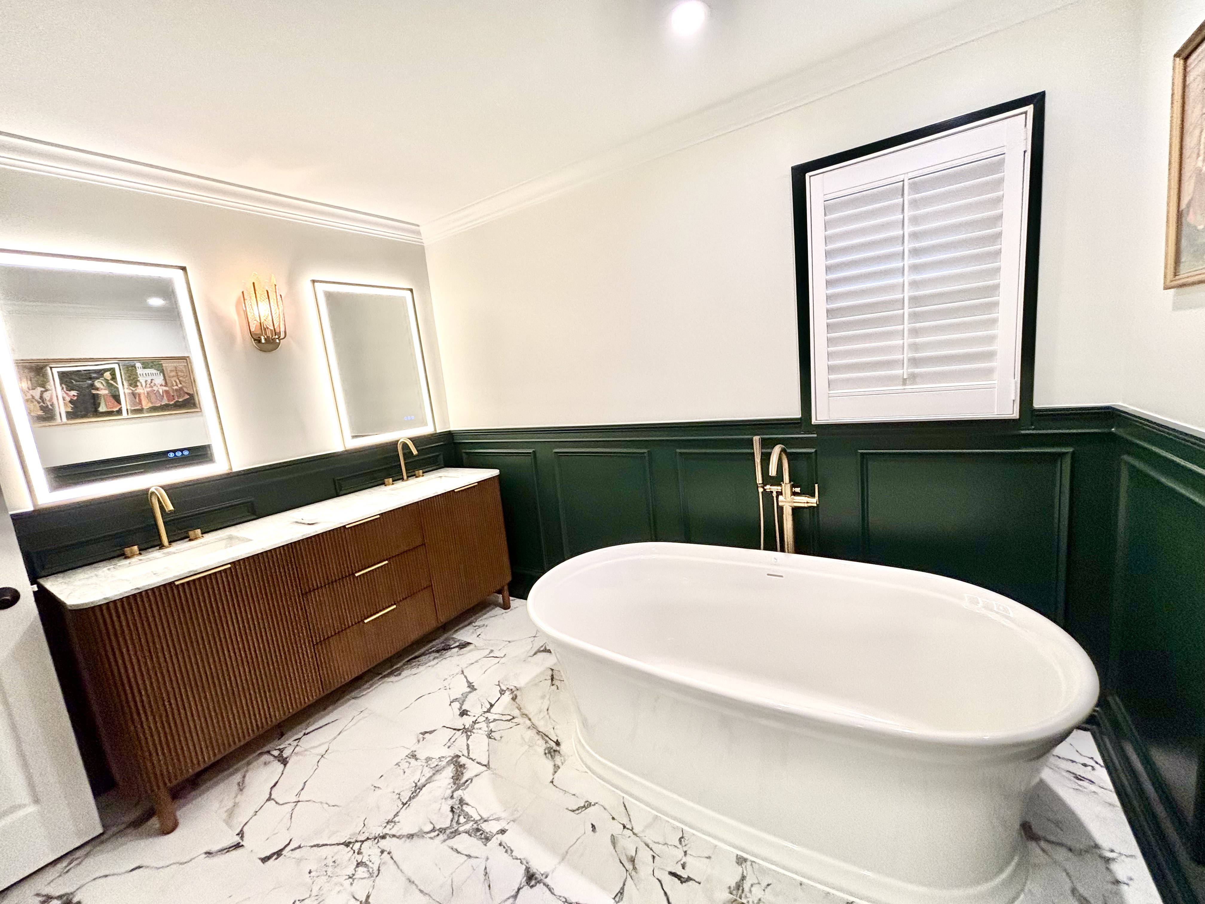 Freestanding soaking tub with a floor-mounted gold tub filler in front of dark green wainscoting and a shuttered window
